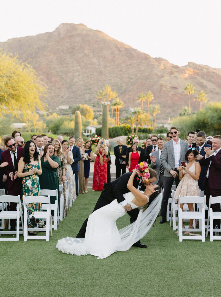 Newlyweds celebrate with a joyful first kiss dip down the ceremony aisle as guests cheer with Camelback Mountain in the background at an Arizona wedding venue.