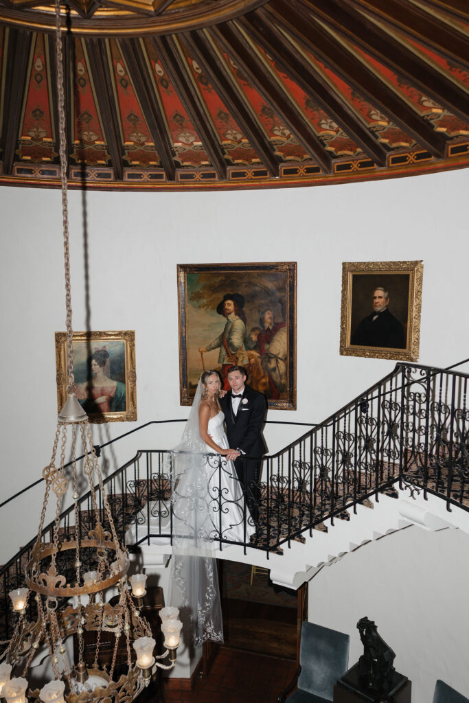 Bride and groom pose on an elegant curved staircase beneath a painted dome and ornate chandelier inside a historic Arizona wedding venue.